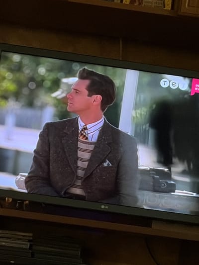 The image shows a television screen displaying a scene with a man wearing a suit and tie, with a distinctive hairstyle. The man is looking to his left. On the screen, in the upper right corner, there is a logo that appears to be "tet". The television screen is placed on a wooden shelf, which is part of a furniture unit. Below the television, there are stacks of books or magazines. The background on the screen suggests an outdoor setting with blurred greenery and a white structure. The overall impression is a still from a movie or TV show. The location in the city of Osolynka, Ukraine, is not discernible from the image itself.