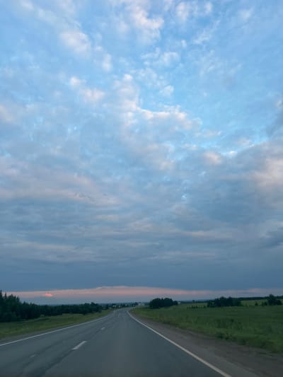 This is a vertical, eye-level, outdoor, daytime, full shot of a paved road curving to the right through a grassy field with trees in the distance, under a cloudy sky. The road is grey asphalt with a white dashed line on the left and a solid white line on the right. The field is lush green grass, and there is a line of dark green trees along the horizon. In the distance, there are small buildings, possibly a village. The sky is a pale blue with scattered white and grey clouds, tinged with pink and orange near the horizon, suggesting it is either sunrise or sunset. The location is identified as Povarёnki, Russia.