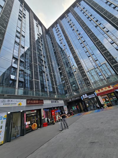 This media file captures a street scene in Linping District, China. The primary subject is a modern, multi-story building with a facade dominated by glass and metal. The building's architecture is characterized by repetitive rows of windows, some of which are open, reflecting the sky. The perspective is tilted, giving a sense of looking up at the building from street level.

On the ground floor, a row of shops with distinct signage lines the street. Visible businesses include a "HAIRSALON" with "发型设计工作室" (Hair Design Studio), a convenience store labeled "嘉沃美便利店" (Jiawo Mei Convenience Store), a restaurant with Chinese characters like "炉火婆婆" (Lu Huo Po Po, likely a restaurant name) and "老挝冰咖啡 泰式奶茶" (Laotian Iced Coffee Thai Milk Tea), and another establishment advertising "手工水餃 过桥米线 重庆酸线粉 豆花饭" (Handmade Dumplings, Crossing the Bridge Noodles, Chongqing Sour Noodles, Tofu Pudding Rice). Some of these storefronts have posters or displays, including an image of a pizza.

Several people are present on the sidewalk. One person is standing with a suitcase, possibly arriving or departing. Others are seen near the shops, some appearing to be customers or employees. A few motorcycles are parked along the sidewalk, suggesting a typical urban environment. The ground is paved with grey tiles. The sky in the reflection of the building appears bright, indicating it is daytime, possibly late morning or early afternoon, with a generally clear or partly cloudy sky. The overall impression is a bustling commercial area in a Chinese city.