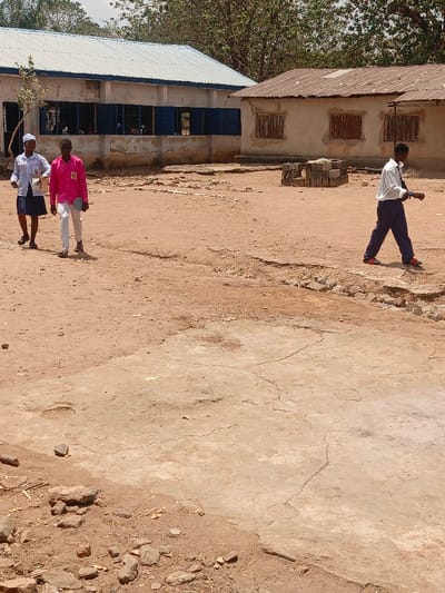 The scene depicts an outdoor area in Mada, Nigeria, characterized by dry, reddish-brown earth and cracked, light-colored concrete patches, indicative of a worn environment. Two single-story buildings stand in the background under a bright, sunny sky.

To the left, a building features light-colored walls, multiple windows with closed blue shutters, and a light blue corrugated metal roof. To its right, an older building exhibits light brown walls, multiple window openings with dark wooden coverings or shutters, and a rusty corrugated metal roof. Several large trees with green foliage are visible behind these structures. A stack of grey concrete blocks is present on the ground between the two buildings, slightly to the right of the center.

Three individuals are walking across the foreground. On the left, two people walk from left to right. The first, a female, wears a white long-sleeved shirt with a blue collar, a dark blue skirt, and a white head covering, carrying a white object in her left hand. The second, a male, wears a bright pink long-sleeved shirt with a white rectangular identification tag on his left shoulder, white trousers, and dark sandals, holding a dark rectangular object in his right hand. Further right and separated from the pair, a third individual, male, walks from left to right. He wears a white long-sleeved shirt, dark blue trousers, and red and white shoes, with a dark object on his left wrist. All individuals are in motion, traversing the open, unpaved ground.