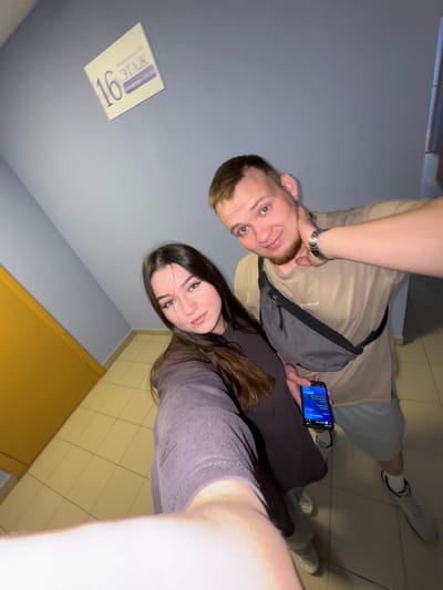 A young couple is taking a selfie in a building hallway with blue walls. The man, on the right, has short brown hair, a beard, and is wearing a beige t-shirt, grey shorts, and white sneakers. He is holding his left hand to his neck and holding a smartphone in his right hand. A grey fanny pack is worn across his chest. The woman, on the left, has long dark brown hair and is wearing a dark grey long-sleeved shirt. She is holding the phone to take the selfie. A sign on the wall behind them indicates "16 ETAL" which translates to "16th Floor." The floor is tiled in a beige color. A yellow door is visible to the left. The overall lighting is dim. The location is identified as Gamovo, Russia, although this is not directly evident from the visual information in the image.