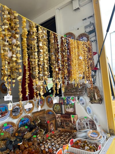 The image depicts a street vendor stall in Baltiysk, Russia, specializing in amber products. Strings of various amber beads, some polished and others rough, hang from a yellow rod, catching the light. These range in color from pale yellow to deep brown. Below the hanging beads, a variety of amber-encrusted items are displayed, including elephant figurines, decorative plates with scenic depictions of Russian landmarks such as St. Basil's Cathedral, and small framed artworks featuring swans and sunflowers. Several price tags are visible, with denominations like "500," "600," "800," "1500," and "200." A yellow truck or van is partially visible on the right side of the frame, suggesting the stall might be mobile. The background shows a white wall and a glimpse of an outdoor environment, likely a street or market area. There are no visible people actively interacting with the stall at this moment.
