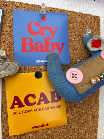 A corkboard displays two square cards and a fabric doll. One card, light blue, reads "Cry Baby" in bold red letters. The other card, yellow, says "ACAB (ALL COPS ARE BASTARDS)" in burgundy. Both include "@Rafaella.clo". A handmade doll, crafted from blue fabric, felt, buttons, and beads, rests on top. A silver keychain and push pins are scattered around the display.