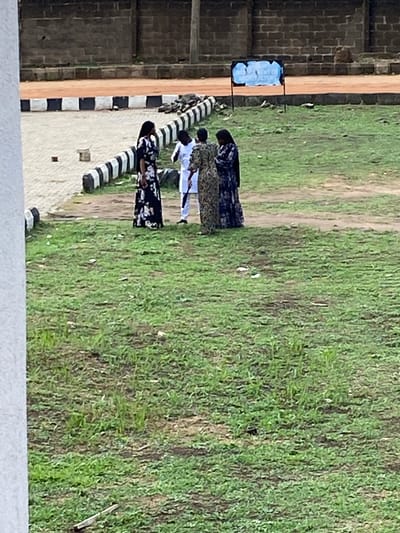 The image depicts a scene in Sabo, Nigeria, showing a group of people in an open, grassy area adjacent to a brick building. Several individuals wear white and black attire and are standing near a black and white striped structure, possibly a barrier or decoration. Other people wearing floral outfits are also present. A sign is visible on the left side of the image. The environment includes grass and dirt.