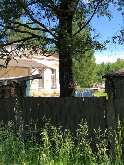 This is a photograph of a building in Izdeshkovo, Russia, on a sunny day. The building is old and appears to be in disrepair, with peeling paint and a weathered roof. It is surrounded by trees and greenery, and there is a wooden fence in the foreground. In the background, there is a stack of lumber and other debris. The sky is blue with a few white clouds.