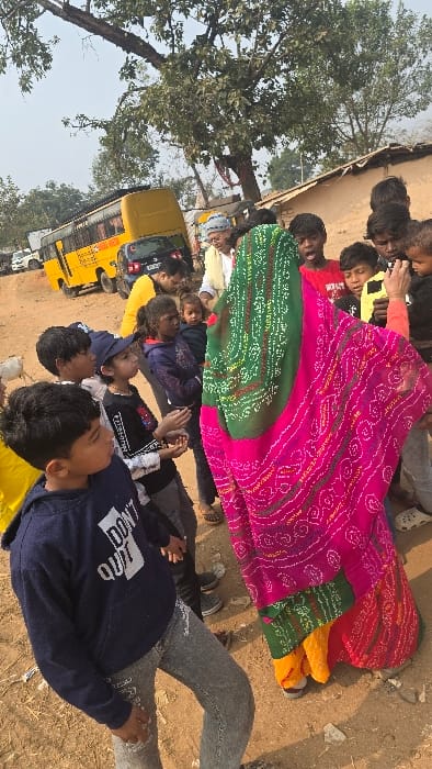 A group lies on the ground, partly covered by a colorful layered cloth. A bus is nearby, indicating a possible school trip. The people are dressed in casual clothes, and a tree is visible in the background, suggesting an outdoor setting. The scene appears to be a moment of rest or relaxation during an event or activity.