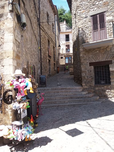 The image depicts a narrow, cobblestone street in Besalú, Spain, characterized by ancient stone buildings on both sides. The street ascends towards the background, with a series of stone steps dominating the mid-ground.

On the left side of the street, a stall displays various stuffed animals, including plush flamingos, unicorns, and ducks, along with a white hat and other trinkets. A sign reading "Carrer Arat d'Oliba" is affixed to the stone wall of a building to the left of the stall. Further up the street, a blackboard sign is visible.

On the right, a stone building features a balcony with dark wooden shutters and a wrought-iron railing. A barred window is also visible on this building. In the background, two young boys are walking up the steps. One is wearing a green t-shirt and shorts, and the other is wearing a blue t-shirt and shorts. Sunlight casts shadows across the cobblestone street and parts of the buildings. The overall atmosphere suggests a historical European town with a lively tourist presence.