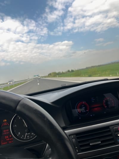 The image captures the view from inside a moving vehicle, likely a car, on a roadway. Through the windshield, a clear, blue sky filled with white clouds is visible, along with a strip of green landscape alongside the road. In the foreground, the dashboard and steering wheel of the vehicle are prominent. The digital display on the dashboard indicates a speed of 148 km/h and a temperature of 13.5°C. The gear is in D6. A warning light is illuminated. The odometer reading is 268835 and the remaining mileage is 182.9 km. The time shown is 09:42.
Given the information, it is not possible to pinpoint the exact location of the scene in Zagore, Bulgaria without further context.