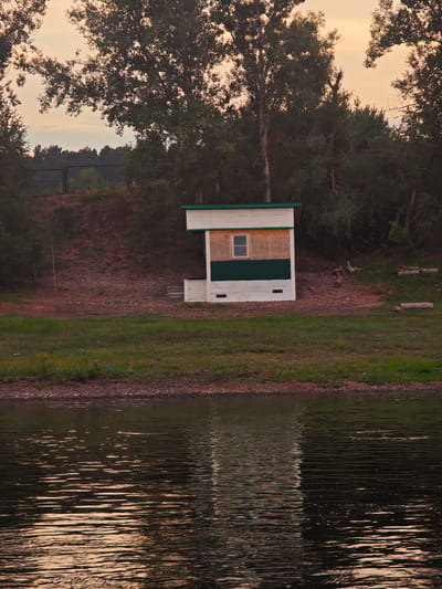The image depicts a small, white and green shed with a single window, situated on a grassy embankment. The shed is positioned next to a slope covered with dry leaves and some scattered logs. Beyond the shed, a dense line of trees forms the background, with a fence visible on a higher level of the embankment. In the foreground, a body of water with rippling reflections of the sky and surrounding foliage occupies the lower half of the frame. The sky is a soft, hazy orange, suggesting either dawn or dusk. No people or animals are present in the scene. The location is identified as Yukseevo, Russia.