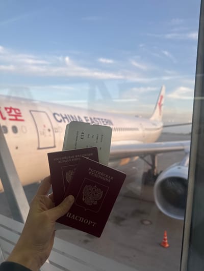 Here's a detailed description of the image:
**General Description:** The image shows a person holding two Russian passports and a boarding pass in front of an airplane window. The aircraft visible outside is a China Eastern Airlines plane.
**Specific Details:**
* **Passports:** Two maroon-colored Russian passports are visible. The inscription "РОССИЙСКАЯ ФЕДЕРАЦИЯ" (RUSSIAN FEDERATION) is clearly visible on the cover of one, along with the word "ПАСПОРТ" (PASSPORT).
* **Boarding Pass:** A boarding pass is partially visible, showing details such as the flight number "MU5066", the date "15MAY26", departure airport "PVG" (Shanghai Pudong), the destination airport, a seat number "41J", and the name "POSTNOVA/EKATE".
* **Aircraft:** The exterior of a China Eastern Airlines aircraft is visible through the window. The airline's name and logo are clearly displayed on the fuselage.
* **Environment:** The scene appears to be inside an airport terminal, near a gate. The window suggests the person is inside the terminal, looking out at the plane. Part of the tarmac and possibly some airport equipment are visible outside.
* **Person:** The hand holding the passport and boarding pass belongs to a person whose features are otherwise obscured.
* **Location:** Given the text "POSTNOVA/EKATE" on the boarding pass, it's possible that the destination is in the Ekaterinburg area. However, the origin point is clearly Shanghai Pudong airport. It's not possible to state that the location is in Bol'shie Kabany, Russia, as that does not appear to be a location related to the details in the photo.