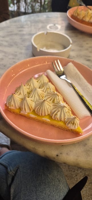 A correctly oriented, bright close-up shot captures a slice of lemon meringue pie served on a pastel pink, heart-shaped plate. The pie, centrally placed, features a bright yellow filling beneath a tall, artfully piped meringue topping, which is lightly torched to a golden brown at its peaks. A silver fork rests on a folded white napkin to the left of the pie.
The setting appears to be a cafe or restaurant table in Bir Mourad Rais, Algeria, as indicated by the location context. The table surface is light-colored, possibly marble or granite, with visible speckles or textures. In the blurred background, a light-colored ceramic cup with a handle is visible to the left, alongside a glimpse of another similar pink plate, suggesting multiple patrons or items. A person's leg, clad in blue denim jeans, is visible in the lower right corner, confirming someone is seated at the table, with a dark object next to them. The natural, bright lighting indicates it is daytime. The overall scene conveys a relaxed and enjoyable moment, likely centered around a dessert break. No text is visible in the image.