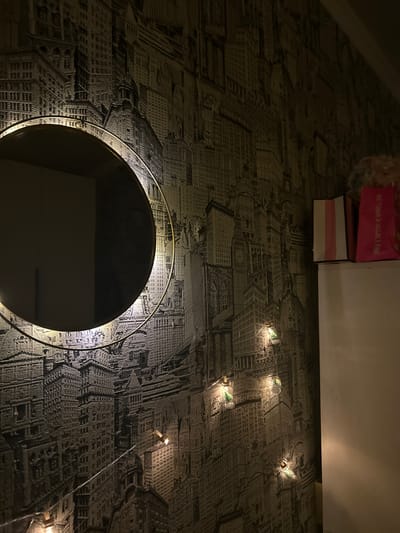 An indoor scene in the city of Raduga, Russia, is depicted in dim, ambient light. The wall is covered with monochromatic wallpaper featuring an intricate and dense cityscape design. A circular mirror with a thin gold-toned frame is mounted on the wall in the left-center, internally illuminated, casting a warm light halo onto the surrounding wallpaper. The mirror reflects a dark, unidentifiable area. A string of decorative fairy lights, composed of small, transparent enclosures containing miniature green tree-like figures, is strung horizontally across the lower and middle sections of the wallpaper, emitting a warm yellow glow. To the right, a light-colored, vertical furniture piece, possibly a cabinet or dresser, stands against the wall. Resting on top of this furniture piece are two retail shopping bags: one white with vertical pink stripes, and another predominantly pink bag displaying the "VICTORIA'S SECRET PINK" logo in white text. A light-colored, possibly fluffy, object is partially visible behind the bags. No individuals or direct interactions are present.