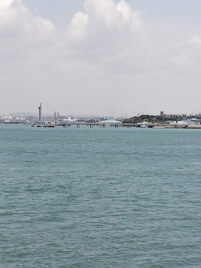 The image displays a wide view of a marine environment looking towards the distant shoreline of Mkomani ward, Kenya, under a mostly overcast sky. The foreground and midground are comprised of a large expanse of teal-blue water exhibiting a rippled, choppy surface texture. In the background, a diverse urban and industrial landscape spans the horizon. From the left, the scene reveals several large, light-colored storage tanks, a tall, slender tower (likely a lighthouse or observation tower), and multiple industrial smokestacks emitting dark plumes. Numerous large ships or vessels are visible in the water adjacent to these industrial facilities. A multi-segment pier or jetty, supported by pilings, extends from the right mid-ground towards the center, featuring a structure with a light-colored roof. Further right along the distant shoreline, additional industrial buildings, port cranes, and a distinct white vessel are present. The far right background shows a cluster of low-rise buildings with darker roofs, surrounded by green vegetation. No individuals are visible in the scene. The sky is dominated by white and grey clouds, indicating diffuse lighting conditions.