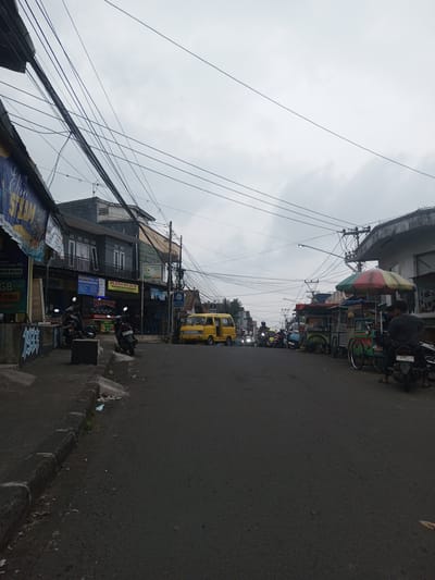 A wide-angle, eye-level photograph depicts a bustling street scene, stated to be in the city of Houaythat, Laos, under an overcast sky. A dense network of black electrical or communication wires spans across the upper portion of the frame.
The asphalt road, dark and appearing somewhat wet, extends into the distance, occupied by a yellow minivan traveling towards the viewer in the middle ground, and numerous motorcycles/scooters, some with illuminated headlights, forming moderate traffic.
On the left side of the street, a concrete curb separates the roadway from a sidewalk. Two motorcycles are parked on the sidewalk, one displaying a license plate 'D 3491 ZM 02 26'. Behind them, a two-story building features a dark gray facade, white window frames, and balconies. Multiple business signs are affixed to this building, including banners with text such as 'WAHID PULSA 24 JAM', 'RINNAY', and 'MASAKAN PADANG'. A blue banner with partially visible white text, including "STLAM," hangs from the building's lower level.
On the right side of the street, several street vendor carts are parked, one prominently featuring a multi-colored umbrella (red, yellow, green). A male individual wearing a dark shirt and sandals is seated on a black motorcycle, license plate 'F 2907 ZM 06-20', with his back to the camera. Another person is partially visible near the vendor carts. Further down the street on the right, a light-colored building is visible with an 'ATM' sign. Litter is scattered along the base of the left curb.