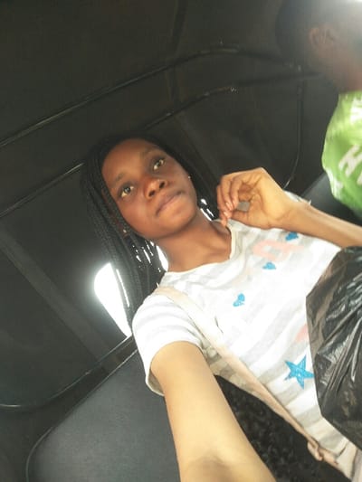An image captured in Ajah, Nigeria, depicts a young female individual seated inside what appears to be a public transport vehicle, likely a tricycle taxi (keke). The primary subject, positioned in the foreground, has dark skin, dark braided hair, and is looking directly at the camera. She is wearing a white t-shirt with thin grey horizontal stripes, accented with blue heart and star motifs. Her left hand is raised, with fingers positioned near her right shoulder/collarbone area. A light-colored strap, possibly from a bag, crosses her chest. To her right, partially visible, is the back and shoulder of another person wearing a green garment. A black plastic bag is visible on the primary subject's lap or adjacent to her. The vehicle's interior features dark, possibly padded, seating and a dark overhead canopy. Bright ambient light enters from an unseen window or opening on the upper left side of the frame.