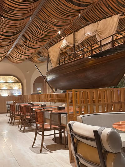 The image depicts the interior of a restaurant with a nautical theme. A large, wooden ship model is suspended from the ceiling, which is adorned with a unique, woven canopy of wooden strips. The ship has fabric sails that are draped and tied. In the foreground and midground, there are rows of dining tables and chairs. The tables have dark tabletops, and the chairs are made of wood with woven seats and backs, or upholstered in a light color. Some of the seating consists of banquettes. The walls are painted in a neutral tone, with arched doorways visible in the background. The flooring is a light-colored, polished tile. The lighting appears to be ambient and spotlights illuminate the ceiling structure. There are no people visible in the image. The style suggests a modern restaurant with a distinctive decorative element. The location provided, Çolpon-Ata, Kyrgyzstan, is not visually evident from the image itself.