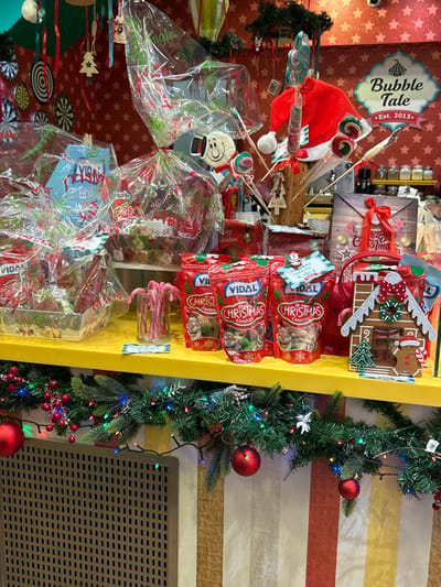 This image captures a vibrant, Christmas-themed retail display within an indoor setting. The scene showcases an array of holiday merchandise and decorations, likely in a shop or market stall.
A prominent yellow display shelf, positioned centrally, holds numerous festive items. On this shelf, several red bags labeled "Vidal Christmas Candies" are stacked. Adjacent to these are various gift-wrapped packages in clear plastic, tied with red ribbons, containing lollipops and other sweets. A gingerbread house-shaped decoration, a red Santa hat, and a small container filled with candy canes are also part of the display. Below the main shelf, more wrapped gifts and circular lollipops are arranged.
To the left of the shelf, a blue sign with "Bubble Tale Est. 2013" written in white is affixed to a red wall adorned with white star patterns. On the right side of the image, a green Christmas garland, strung with multicolored lights, red baubles, and red berries, hangs against a striped wall featuring alternating bands of brown, yellow, and red. A black vent or grille is visible at the bottom right corner of this wall.
The visible text includes "Vidal Christmas Candies" on the red product bags, "Merry Christmas" on a gift tag, and "Bubble Tale Est. 2013" on the overhead sign. The overall impression is one of festive holiday cheer and an abundance of seasonal treats and decorations available for purchase.