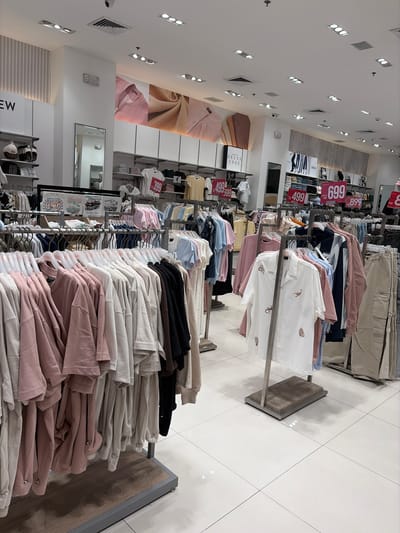 This media file shows the interior of a clothing store in Dagupan, Philippines. Racks of clothing, primarily shirts and blouses, are neatly displayed on metal racks. The shirts are in a variety of colors, including white, pink, blue, beige, and black. Some shirts have price tags indicating prices such as 399, 499, 699, and 899 PHP. The store appears to be well-lit with overhead lights. The flooring is tiled. There are also promotional signs visible with text like "DRESS CODE" and "DENIM". The store seems to be empty of customers, suggesting it might be early in the day or not a peak shopping time. The overall impression is that of a clean and organized retail environment.