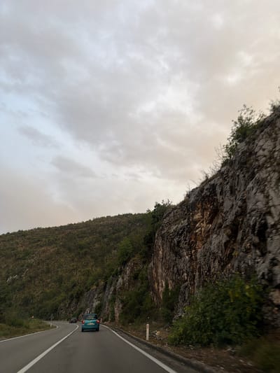 The image depicts a road in Drenoštica, Montenegro, winding through a mountainous landscape under an overcast sky. A turquoise car is traveling on the road, with another dark-colored vehicle in front of it. The road is paved and has white lane markings. To the right of the road, a steep, rocky cliff face rises, covered in patches of green vegetation. To the left, a green, treed hillside slopes upwards. The overall impression is of a scenic drive through a rugged, natural environment.