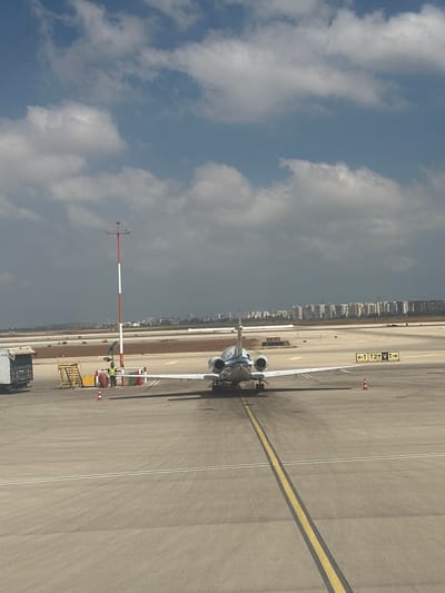 An airplane is parked on a tarmac at Ben Gurion International Airport in Yehud-Monosson, Israel. The airplane is a private jet with a white fuselage and twin engines. The tail of the plane is visible, with the engines mounted on either side. The plane is on a paved surface with yellow and white lines marking the taxiway. In the background, a row of apartment buildings can be seen in the distance, suggesting an urban environment. The sky is partly cloudy, with patches of blue sky visible. There are several airport-related objects visible, including a utility pole with lights, a small set of stairs, and what appears to be a luggage cart. A person wearing a high-visibility vest is standing near the utility pole. The overall scene depicts a typical airport environment on a clear day.