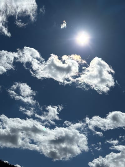 The image captures a bright, sunlit sky with scattered white clouds. The sun is prominently visible as a radiant orb, casting a lens flare. The clouds exhibit varying textures, from wispy cirrus formations to denser cumulus clouds, some tinged with iridescent colors possibly due to atmospheric conditions. The sky is a deep blue. In the bottom left corner, the dark silhouette of a tree line is visible against the sky, suggesting an outdoor, possibly elevated, vantage point. There is no indication of the city of Gela, Bulgaria.