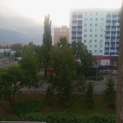 This media file depicts an outdoor scene in Meleuz, Russia, likely taken from an elevated viewpoint. The primary subjects are residential buildings, trees, and a street with a few parked cars.
In the background, a tall, light blue apartment building with blue accents on each floor dominates the skyline. To its left is a shorter, tan-colored apartment building. In front of these buildings, a variety of trees, including tall, slender deciduous trees and some evergreens, are scattered across the landscape.
A street runs through the middle ground, with a few cars visible. One car, a light-colored sedan, is parked with its trunk open. Another similar car is parked nearby. A blue delivery truck is also visible on the street. A small strip of green grass and a decorative black metal fence separate the street from the area with more dense foliage in the foreground.
In the foreground, there are more trees and bushes, with thick green foliage. The overall impression is that of a residential area with a mix of urban structures and natural elements.
The time of day appears to be late afternoon or early evening, given the diffused light and the slightly overcast or hazy sky. The weather seems to be mild, with no obvious signs of rain or strong sunlight.
There is some text visible on the facade of a building adjacent to the street. It appears to be a store or commercial establishment. One sign has a red and white striped design with a logo that looks like a stylized "M". Another sign to the left has smaller text that is difficult to read.
The scene evokes a sense of everyday life in a Russian town, with a calm and somewhat muted atmosphere. There are no people prominently featured, emphasizing the setting itself.