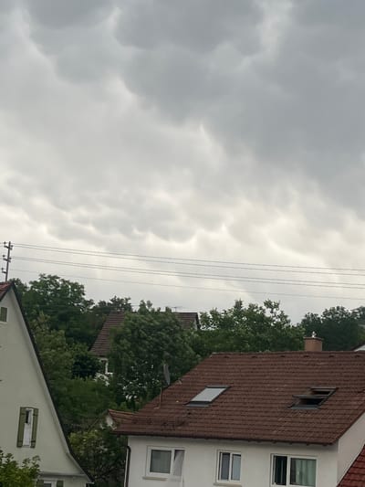 The image depicts a cloudy sky with mammatus clouds, indicative of an approaching storm, above a residential area in Balingen, Germany. In the foreground, the roof of a house with red tiles is visible, featuring two skylights, one open. Another house with a light-colored facade and green shutters is partially visible on the left. Electrical wires are strung between poles in the middle ground, with lush green trees obscuring some of the houses behind them. The overall scene suggests a somber, overcast day.