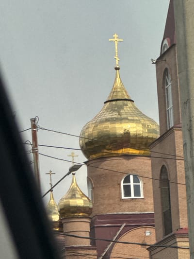 This media file shows the exterior of a building with distinctive golden domes, characteristic of Orthodox churches. The building appears to be constructed with brickwork, featuring several windows of various shapes, including arched and rectangular ones. One notable detail is a distinct arched window with a white frame, divided by a cross-like structure. Several golden crosses are visible atop the domes.
The image is taken from a low angle, looking upwards towards the building. Several dark wires or cables crisscross the frame, obscuring parts of the building and the sky. The sky is overcast and a pale grey, suggesting a cloudy day. There are no people visible in the image.
The overall impression is of a religious structure in an urban or semi-urban setting. The golden domes are the most prominent feature, reflecting the light and drawing attention to their intricate, faceted surfaces. The presence of crosses further reinforces the religious context. The weather appears to be cool and perhaps damp, given the overcast sky. The view appears to be from ground level, possibly from across a street or open area. No text is visible in the image. The image has been rotated 90 degrees clockwise.