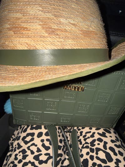 The image shows a close-up view of a straw hat with a green leather band. Below the hat is a green clutch bag with an embossed "Steve Madden" logo repeated in a grid pattern. A piece of leopard-print fabric is visible at the bottom of the image, possibly part of an outfit or another accessory. The overall scene appears to be a flat lay or a still life arrangement. The lighting suggests an indoor setting, and the background is dark and out of focus. There are no people or discernible locations in the city of Kennesaw, United States of America.