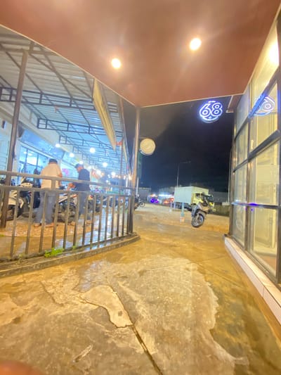 This image, presented sideways as if rotated 90 degrees clockwise, captures a vibrant night scene in Keudeublang, Indonesia, with visibly wet ground suggesting recent rain.
To the left (which would be the top if rotated vertically), a covered outdoor area features a brown ceiling with bright light fixtures illuminating the space. Above, glass panels reflect light. A prominent blue neon sign, shaped like a circle, displays the number "89" and the text "COME JOIN US". Below it, a smaller circular sign is partially visible, showing "CLINIC" and "BOBBY".
Underneath this canopy, a black metal railing separates the space from the foreground. Several motorbikes are parked in this area. At least two men are visible near the railing and motorbikes; one wears a dark shirt, the other a white shirt, both engaged in indistinct activities, possibly conversing or preparing to leave. More figures can be seen deeper within the covered area, which appears to be a cafe or gathering spot with tables and lights.
The immediate foreground and extending to the right (the bottom if rotated vertically) show a cracked concrete or paved ground, extensively wet with reflective puddles and damp patches. Small sprigs of green grass grow along the base of the metal railing. Beyond the railing, under the dark night sky, streetlights glow in the distance. A motor-scooter is parked on the wet ground, and further down the street, a light-colored box truck is visible. The numerous artificial lights scattered across the scene create a lively, urban atmosphere at night.