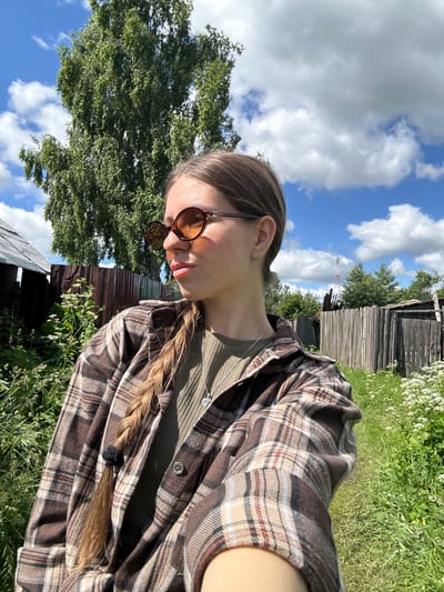 In this medium shot, a young woman with long, braided brown hair and brown-rimmed sunglasses is pictured from the chest up. She is wearing a brown and beige plaid shirt over a green ribbed t-shirt and a silver heart pendant necklace. The woman is outdoors in what appears to be a rural setting. A large green tree stands to her left, and a wooden fence stretches across the background. The sky is blue with scattered white clouds. The overall scene suggests a sunny day. The location is in the village of Malaya Borshchevka, Russia.