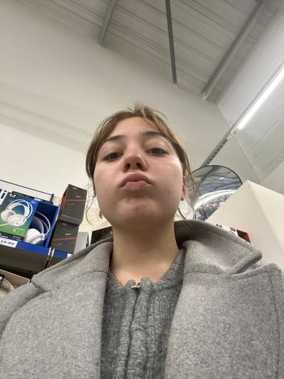 A young woman looks down at the camera with a pout. She has light brown hair and is wearing a gray jacket. Behind her, there is a ceiling with fluorescent lights, a cooling fan, and shelves stocked with headphones. Some of the headphones are visible in their packaging. The image is slightly tilted.