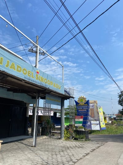 The image shows a street corner in Kecamatan Tulangan, Indonesia (latitude: -7.484, longitude: 112.652). There is a blue and yellow sign for a restaurant called "Jadoel Singop ABU" which is a Mie Pedos & Pinsur shop. To the right is a sign for a school,  SMAN 1 TULANGAN, with the words "SMA NEGERI 1 TULANGAN - SURABAYA - JAWA TIMUR" printed on it. There are a few small trees and shrubs in the foreground. The ground is paved with concrete blocks. The sky is blue and sunny.  There are power lines running across the street, creating a network of black wires against the bright sky.