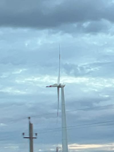 The image captures a wind turbine with a distinct red and white striped blade against a cloudy sky. In the foreground, a power pole with visible wires is present. The overall scene appears to be an outdoor industrial or rural setting, possibly on the outskirts of the city of Zimovniki, Russia, given the presence of wind turbines, which are common in such areas. The lighting suggests it might be overcast or dusk.