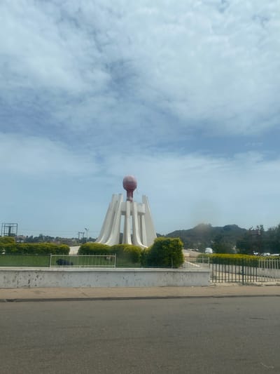 The image shows a monument in Ladura Foron, Nigeria. The monument consists of a tall, white, curved structure with several vertical supports, topped by a large, reddish-brown sphere. It is surrounded by manicured green bushes and a white concrete wall with a black metal fence. In the background, there are hills covered with vegetation, and a cloudy sky. In front of the monument, a paved road is visible. There are no people in the image, but two dogs are lying on the grass within the fenced area.
