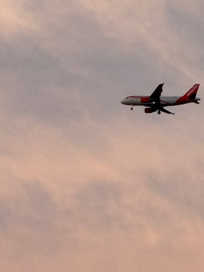 An EasyJet airplane is captured in mid-flight against a dusky sky. The aircraft is predominantly white with orange accents and the "easyJet" logo clearly visible on its tail. The sky is a gradient of soft purples and pinks, with visible cloud formations. The airplane appears to be on its descent, with landing gear extended, suggesting it is approaching an airport. The image is framed with the airplane positioned in the upper right quadrant, leaving a significant portion of the sky empty in the lower left. This scene was photographed in the vicinity of Grenay, France, at an unspecified time, likely during twilight.