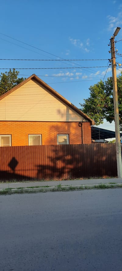 The image captures a side view of a two-story building with a distinct architectural style. The lower part of the building is clad in reddish-brown brick, while the upper, triangular gable section is covered in light-colored vertical siding. Three rectangular windows are visible on the brick portion of the building, and one on the siding. The house is situated next to a paved road, with a strip of grass and weeds separating the road from the building.
To the left of the building, a utility pole with multiple wires extends upwards, disappearing into the frame. A large, leafy green tree is positioned behind and to the left of the house, partially obscuring the roofline. The sky is a clear, vibrant blue with a few scattered white clouds.
The lighting suggests it is late afternoon or early evening, with the sun casting long shadows from unseen foliage onto the wooden siding of the building. The shadows create an abstract, dappled pattern on the wall, adding depth and texture to the scene. The overall impression is one of a residential or perhaps a small commercial building in a semi-urban or suburban environment. The image is oriented vertically, with the ground appearing on the right side of the frame and the sky on the left. No text is visible in the image.