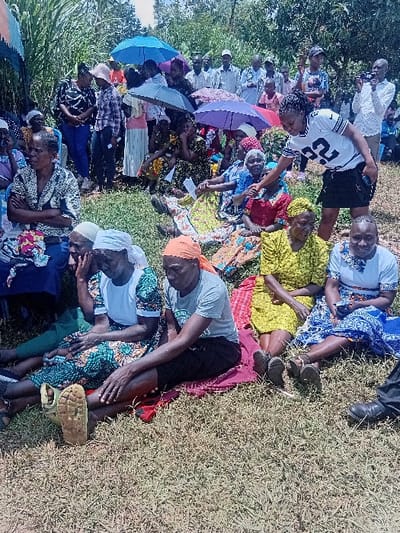 An outdoor gathering of approximately 40-50 individuals is documented in Butali Chegulo ward, Kenya, under bright daylight conditions. The scene is set on dry, sparse grassy terrain. In the foreground, numerous individuals, primarily elderly women, are seated directly on the ground, some utilizing red and blue fabric mats. Their attire includes various patterned dresses, shirts, and head coverings. A young woman, identified by a white and black jersey with "32" emblazoned on it and a black skirt, stands in the mid-ground, extending her right hand towards an older, seated woman. Behind this interaction, a larger group of men and women are visible, both standing and seated, with several holding open umbrellas (blue, purple, grey, black) for shade. Dense green foliage forms the background. On the right side, a person is observed holding a camera or smartphone towards the assembled group. A segment of a blue and orange striped canopy is visible on the far left. Individuals display varied states of attention, with some observing the central interaction.