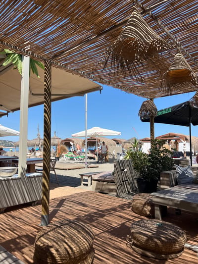 The image depicts a beach bar in San Roque, Spain, on a sunny day. The foreground is a wooden floor covered with a woven mat, with two large, circular, woven poufs casting shadows from the overhead thatched roof. Beyond the poufs, a wooden table and bench are visible. A large wooden pillar, part of the beach bar's structure, stands prominently, also casting striped shadows.
In the middle ground, the beach is populated with thatched umbrellas and sun loungers, some occupied by people. A white umbrella with "Cruzcampo" branding is visible. There are structures with thatched roofs that appear to be part of the beach bar's seating areas. A black umbrella with a "Heineken" logo is also present. Green plants are placed in pots.
The background shows the clear blue sea and the horizon under a bright blue sky. The overall scene suggests a relaxed atmosphere at a beachside establishment.