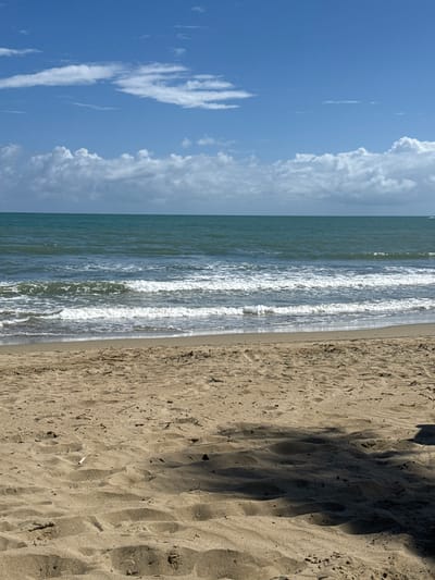 The image depicts a beach in Cabarete, Dominican Republic. The scene is divided into three main sections. On the left, a blue sky is filled with cumulus clouds. The middle section features the ocean with gentle waves breaking onto the shore. The right section shows the sandy beach with visible footprints and shadows cast by trees or structures located off-frame. There are no people visible in the image.