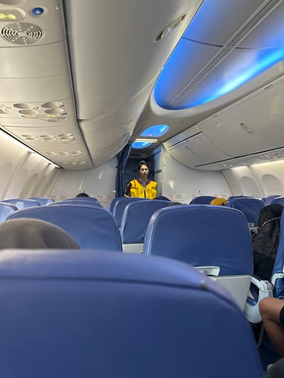 The image depicts the interior of an airplane cabin, likely on a commercial flight. In the foreground, rows of empty blue airplane seats are visible, some with headrests and seatbelt buckles. The perspective suggests the photo was taken from a passenger's seat. In the middle ground, a flight attendant is standing in the aisle, facing forward, wearing a yellow vest over a uniform. Her posture and attire indicate she is on duty. The cabin ceiling features overhead compartments, air vents, and mood lighting in a striking blue hue, creating a modern ambiance. The overall environment is orderly and typical of an aircraft cabin. The provided location of Baji Mangngai, Indonesia, suggests this is likely a flight operating within or to that region.