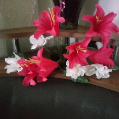 The image displays a floral arrangement composed of bright pink and white artificial flowers. The flowers are arranged on a wooden shelf, with some foliage visible. In the background, there is a dark vase or pot, possibly containing the flowers, and a mottled green wall. The lighting suggests an indoor setting, possibly during the day. The overall scene is a still life of decorative flowers. There is no discernible text, people, or activity taking place.