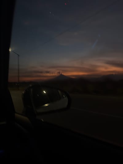 This media file captures a dramatic sky scene from the interior of a vehicle, looking out of the side window. The image is oriented horizontally, depicting a dynamic cloudscape during either dusk or dawn. The sky is dominated by large, dark, and textured clouds, which are intensely illuminated by a warm, orange-brown light, creating a strong contrast with the darker grey and black areas of the sky. This illumination gives the clouds a fiery, almost painterly appearance.

In the lower right portion of the frame, the black side-view mirror of the car is prominent. It reflects a bright, streaked light source, indicative of either artificial street lighting or headlights from another vehicle. The car's interior, specifically the window frame and a section of the dashboard, is visible along the bottom and right edges, partially framing the view. Subtle reflections or lens flares can be observed on the window surface, particularly in the darker upper-left quadrant. The overall impression suggests motion, capturing a fleeting moment of striking natural light from within a vehicle. No visible text is present in the image.