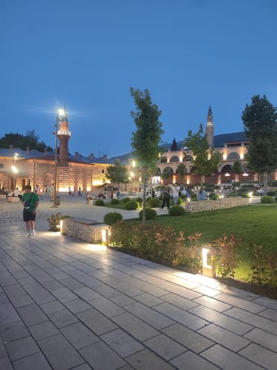 An evening view of the Osmangazi district in Bursa, Turkey, showcases a courtyard area illuminated by artificial lights. A young male wearing a green t-shirt and dark shorts walks on a paved walkway in the foreground, facing left. Behind him, a stone building with a minaret is lit up, and several people are seated and standing in the courtyard. The scene includes trees, neatly trimmed bushes, and a well-maintained lawn, all bathed in the soft glow of pathway lights and architectural lighting, suggesting a public space designed for evening recreation. The sky is a deep blue, indicating dusk or early night.