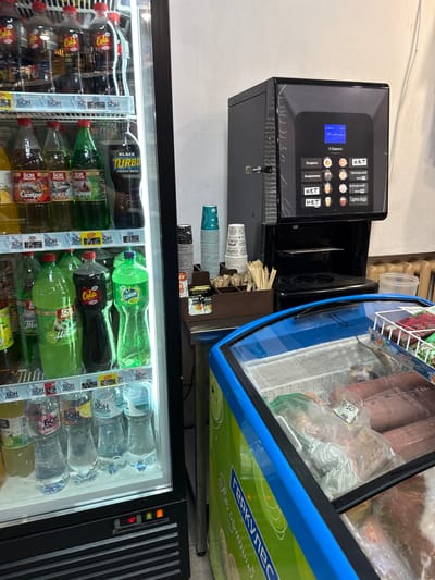 The image captures an indoor commercial setting in Donets, Ukraine, likely a small store or self-service area. The scene features a coffee vending machine, an open-top freezer, and a large upright glass-door refrigerator filled with beverages.
In the upper left, a black Saeco Phedra coffee machine displays selection options on its screen. Visible text includes "Ecпpeco" (Espresso), "Американо" (Americano), "Гаряча вода" (Hot water), and "Ірландський" (Irish), with some options marked "нет" (not available). Beside the machine are stacks of disposable cups, some bearing "COFFEE" and "Greenfield" branding, along with stirrers.
To the right of the coffee machine, an open-top freezer holds various plastic-wrapped frozen food items. A price tag with "-50" and "9889" is visible on one package. The side of this freezer features the text "ГЕРКУЛЕС" (Hercules).
Dominating the lower half of the image is a brightly lit glass-door refrigerator packed with bottled drinks. Various brands and types are discernible, including "Cola", "BLACK TURBO ENERGY DRINK", "LEMON FRESH", "Cola FRESH", "МЯТА" (Mint), and "Ситро" (Citro). The brand "БОН КВАССОН" (BON KVASSON) appears frequently on price tags, which show values like "18.50", "35", "46", and "22.50". The refrigerator's control panel displays a temperature of "4.2". No people are present, depicting a quiet retail setup.