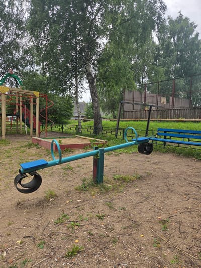This is an outdoor image of a playground in the city of Mishutino, Russia. In the foreground, a turquoise seesaw with blue seats is positioned on a sandy ground with scattered debris. The seesaw is centrally located, with its left end slightly lower than its right end. Behind the seesaw, to the left, is a sandbox with red borders filled with sand. Further back, a colorful playground structure with yellow and red elements is visible. To the right of the seesaw, a blue bench is situated on the grass. The background features a chain-link fence, trees with green foliage, and a cloudy sky. The overall scene depicts a well-used but somewhat dated playground in a residential or park area. There are no people visible in the image.