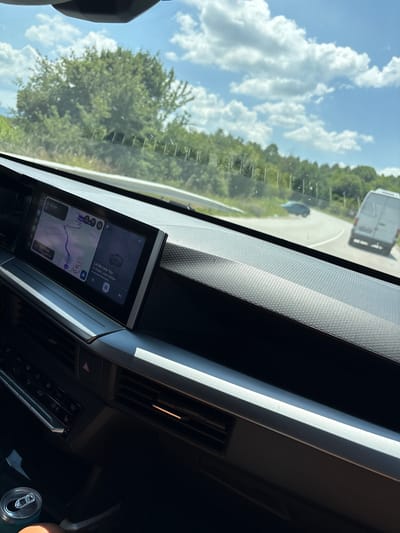 The image shows the interior of a vehicle, likely a car, viewed from the passenger seat. The dashboard features a large touchscreen display showing a navigation map and music playback. Outside the car, a road curves to the right, with a white van and a dark-colored car visible ahead. The landscape is lush and green, with trees and vegetation lining the road under a bright, partly cloudy sky. The overall impression is a snapshot of a road trip through a scenic rural area, possibly near Padarino, Bulgaria.