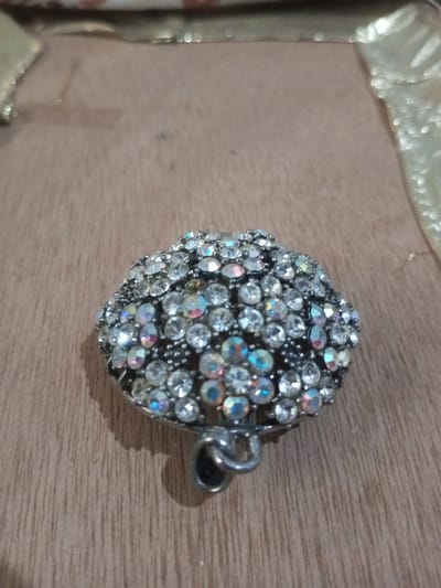 A dome-shaped piece of costume jewelry, heavily encrusted with numerous small, circular, faceted rhinestones, is depicted centrally. Most rhinestones are clear, while some exhibit an iridescent, multi-color shimmer. The base metal of the jewelry appears dark-toned, possibly antique silver or blackened. A metal loop, or bail, for suspension is visible at the bottom of the object. The jewelry rests on a light brown wooden surface displaying a distinct grain pattern. In the blurred background, golden or light-colored fabric or material is partially visible. No individuals, actions, or interactions are visible. The specific location within the city of Chandragiri, Nepal, cannot be determined from the visual information provided in the image.