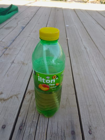 A green plastic bottle of "ilton Tea" with a yellow cap sits on a weathered wooden surface. The bottle is partially filled with a yellowish-green liquid. The label on the bottle features images of peaches and the text "ilton Tea," "500ml," "Shaftoli," "te choiy," and "Nice tea." The wooden surface appears to be a deck or table, with visible grain and nail heads. In the upper left corner, a green cloth is draped. The scene suggests an outdoor setting, possibly during the daytime due to the lighting. The overall impression is casual and relaxed. There are no people visible in the image. The location is identified as Uchqizil, Uzbekistan.