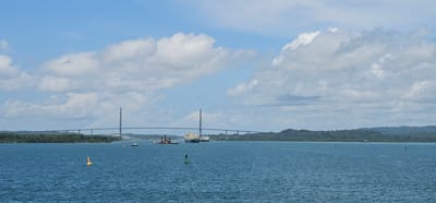 This is a wide-angle, horizontal, exterior, daytime photograph taken from a body of water. The dominant feature is a large, modern, cable-stayed bridge spanning a wide waterway. The bridge has two tall, angular pylons, from which cables extend to support the roadway. The roadway is relatively flat and appears to extend across the entire width of the frame. The bridge is situated in Barrio Norte, Panama, a location known for its proximity to the Panama Canal.
In the foreground, the deep blue water is visible, with several buoys marking the channel. A yellow, conical buoy with a white top is in the mid-left of the frame, and a green cylindrical buoy with a white top is in the mid-right. Further back, in the middle ground, a group of vessels are gathered. These include what appears to be a dredging barge or platform, surrounded by smaller boats. Two large cargo ships are positioned to the right of the dredging equipment.
The background consists of a lush, green, tree-covered coastline with rolling hills. The sky is a bright blue, dotted with scattered, fluffy white clouds. The overall impression is one of a busy, functional waterway and its supporting infrastructure.